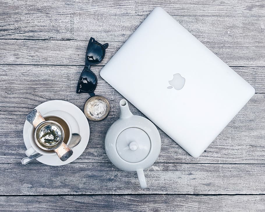 silver MacBook beside white ceramic teapot, high angle photo of silver MacBook, white ceramic teapot, black Ray-Ban sunglasses, and a cup of coffee on brown wooden board