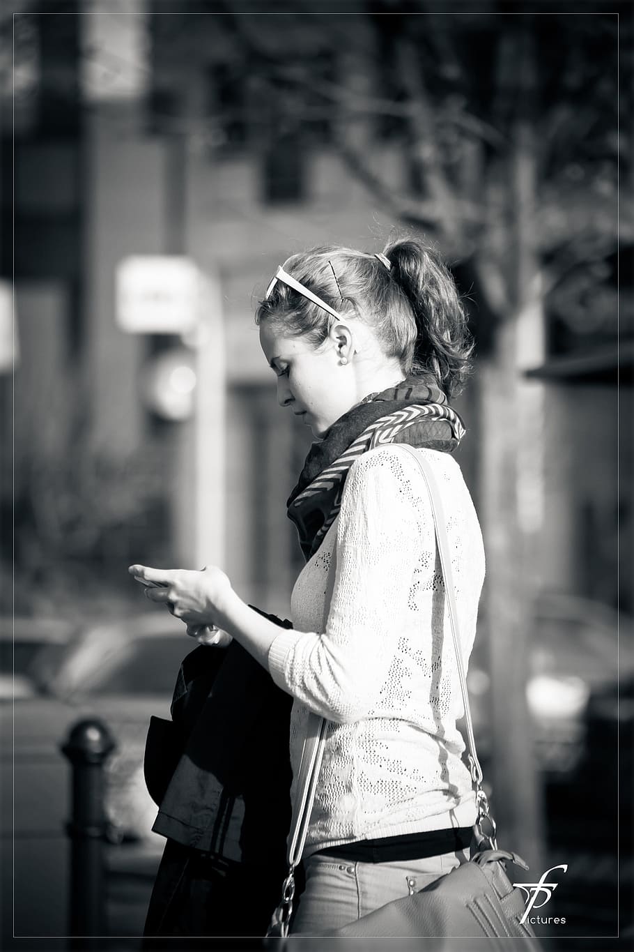 grayscale photography of a woman wearing sweatshirt and scarf walking on street while looking at her phone