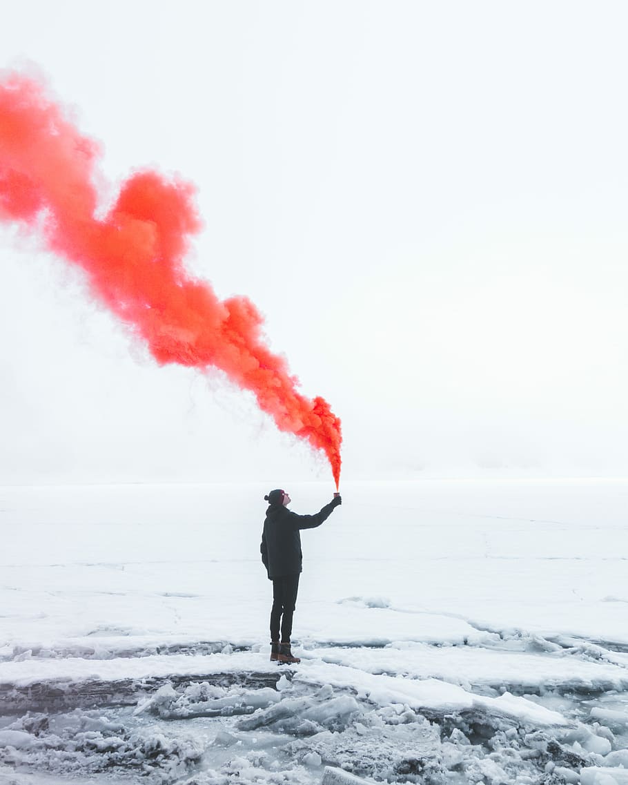 person wearing black jacket and holding red colored smoke flare during daytime, person in black jacket standing on snowy field while holding container with orange smoke, HD wallpaper