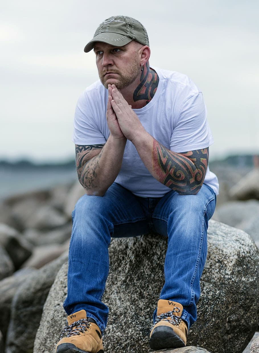 tattooed man wearing gray baseball cap, white crew-neck t-shirt, and blue denim jeans sitting on gray boulder under gray clouds, HD wallpaper