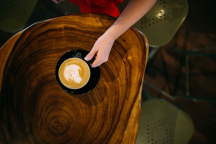 person holding teacup with cappuccino inside on top of wood slab table, high angle photo of person holding a cup of coffee HD wallpaper