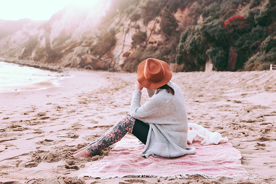 photo of person sitting on sand at seashore, woman sitting on pink shawl near body of water HD wallpaper