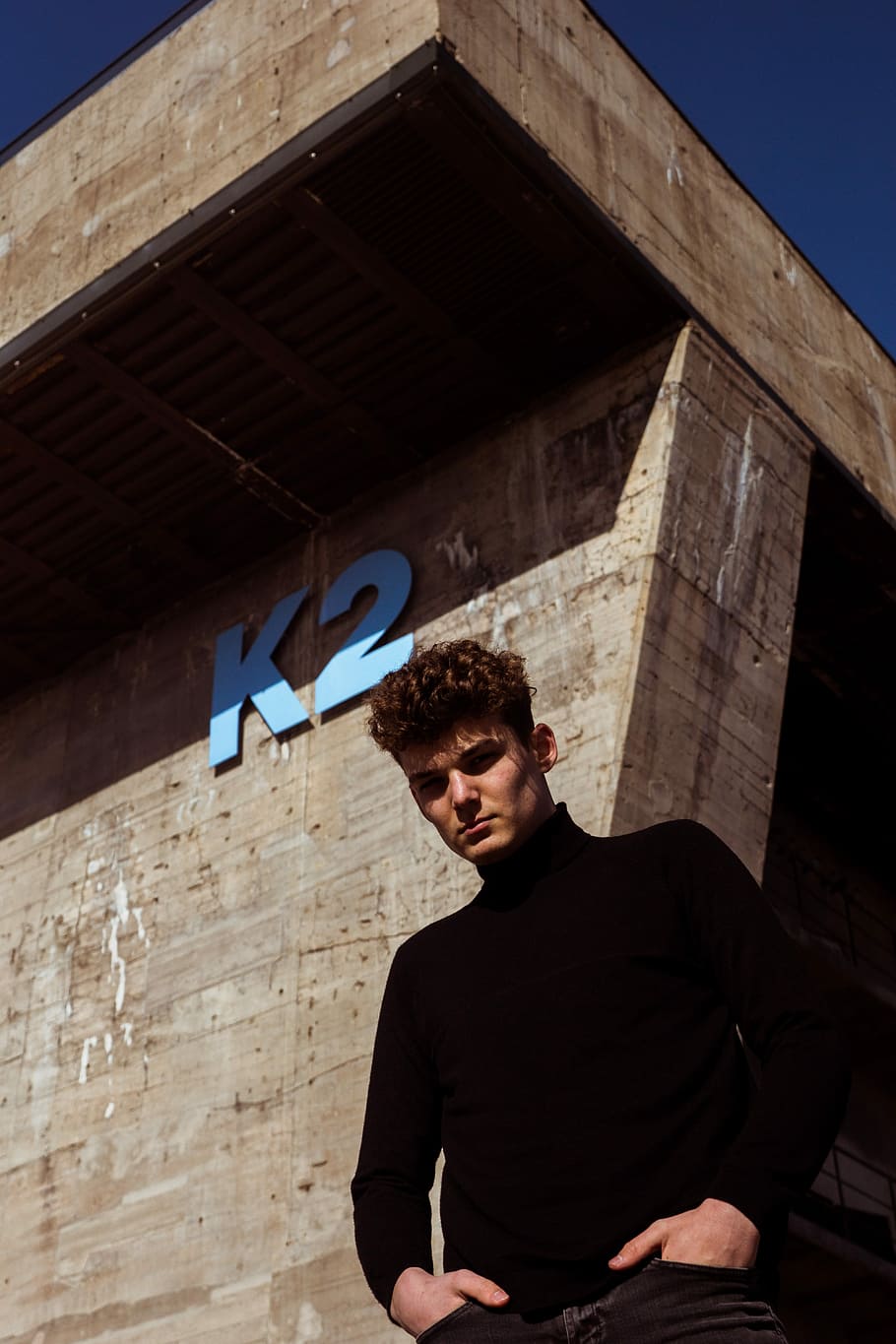 man wearing black turtleneck sweater standing near K2 building, man in black long-sleeved top standing beside gray K2 concrete building HD wallpaper