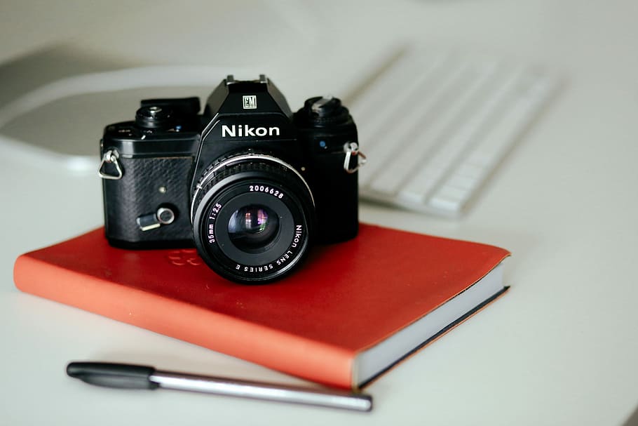 black Nikon MILC camera on red book and pen, black Nikon point-and-shoot camera on brown notebook next to ballpoint pen HD wallpaper