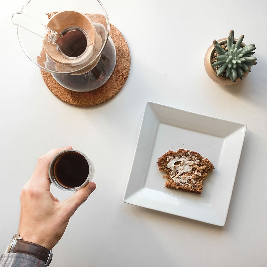 person holding cup of tea beside plate, clear glass teapot on round brown particle board trivet HD wallpaper