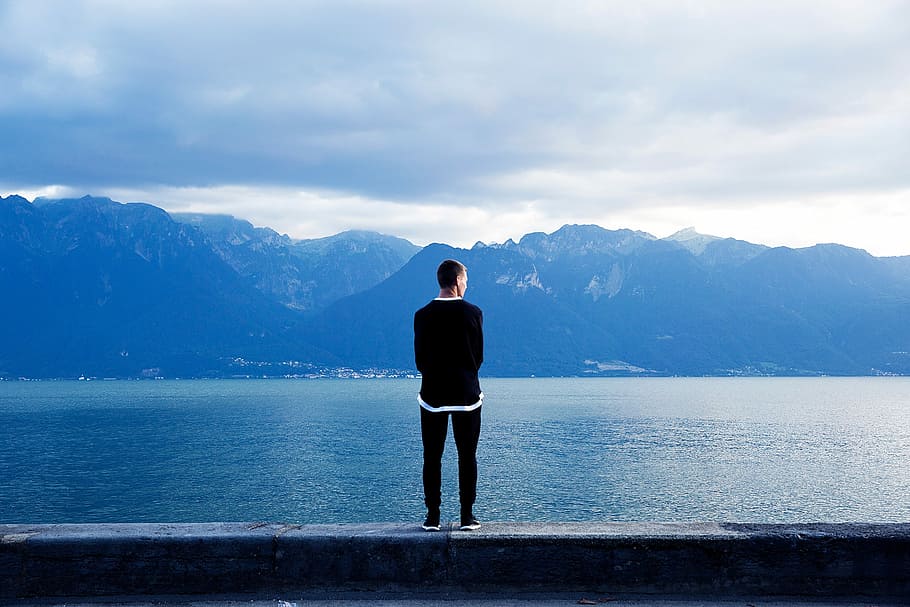 man in black sweater facing the alps across the calm sea during daytime HD wallpaper