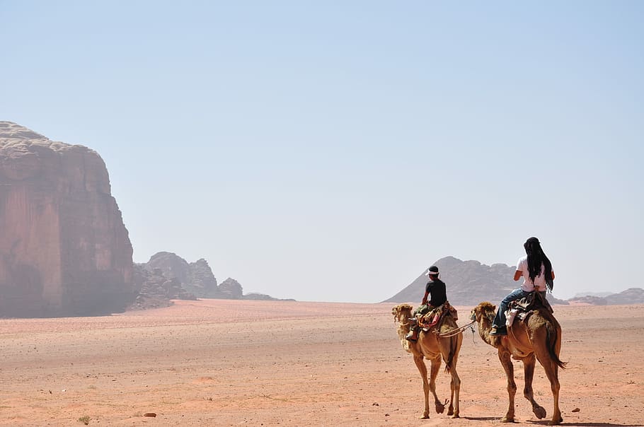 man and woman riding on horse during daytime, two men riding camels walking on desert with view of brown rock formations during daytime photo