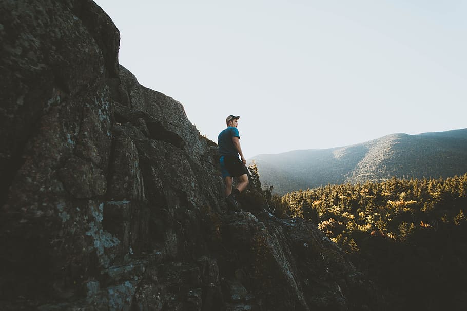 man standing on mountain cliff, man standing on rock formation near forest HD wallpaper