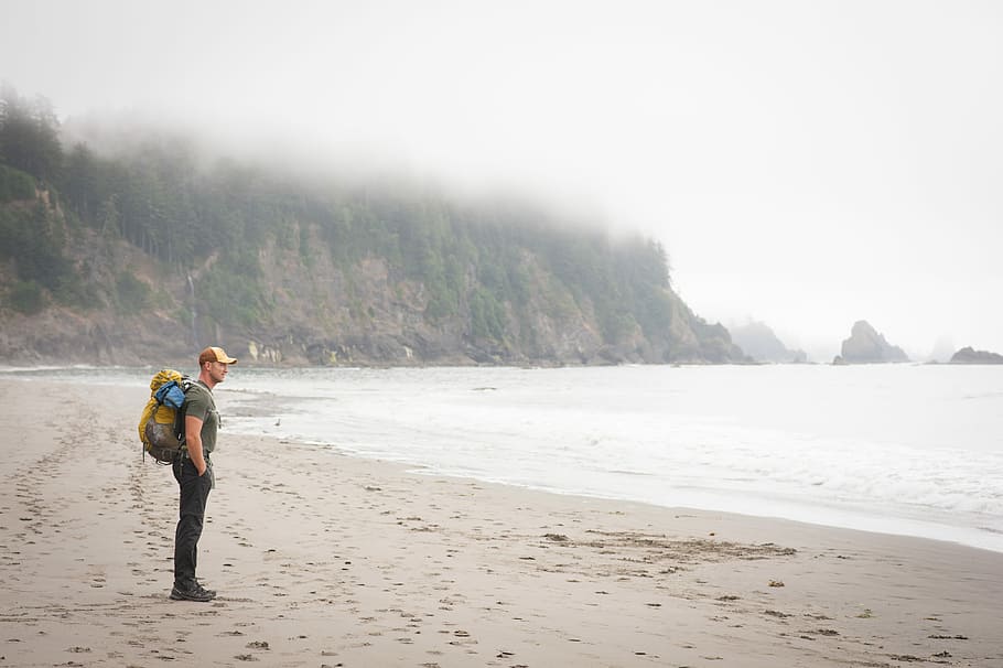 person carrying backpack standing on sea shore, man standing on shore near ocean during daytime HD wallpaper