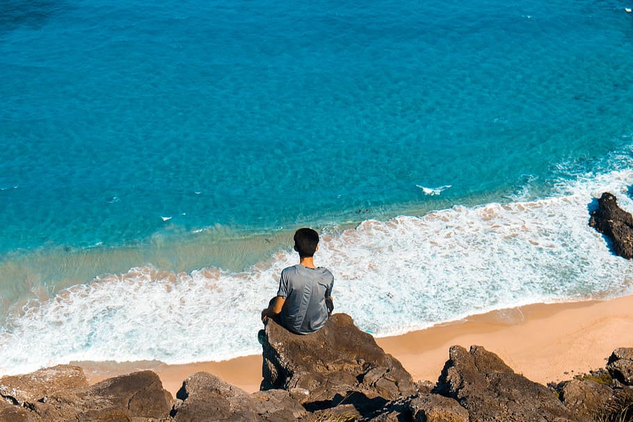 man sitting on top of cliff while looking on seashore, man wearing gray shirt sits on cliff in front of sea during daytime HD wallpaper