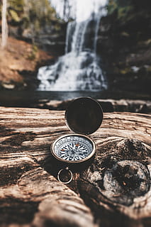 brass-colored compass on brown wooden tree stump in front of waterfalls during daytime, depth of field photography of navigational compass HD wallpaper
