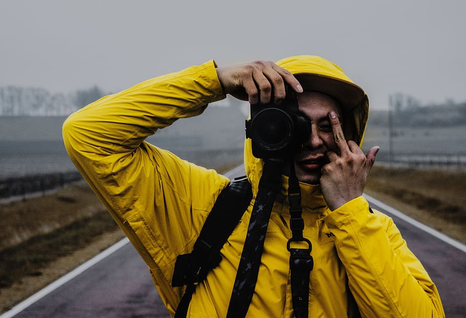 man holding black DSLR camera and raising middle finger standing on concrete road at daytime, person in yellow hooded jacket holding camera
