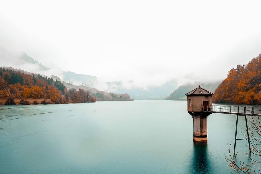 white lighthouse with brown bridge surrounded with body of water under white clouds, beige concrete gazebo dock surrounded by mountains and forest HD wallpaper