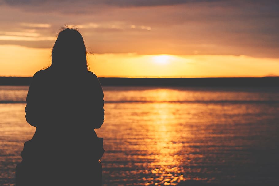 silhouette of woman sitting near bay, silhouette of person sitting facing towards body of water during golden time