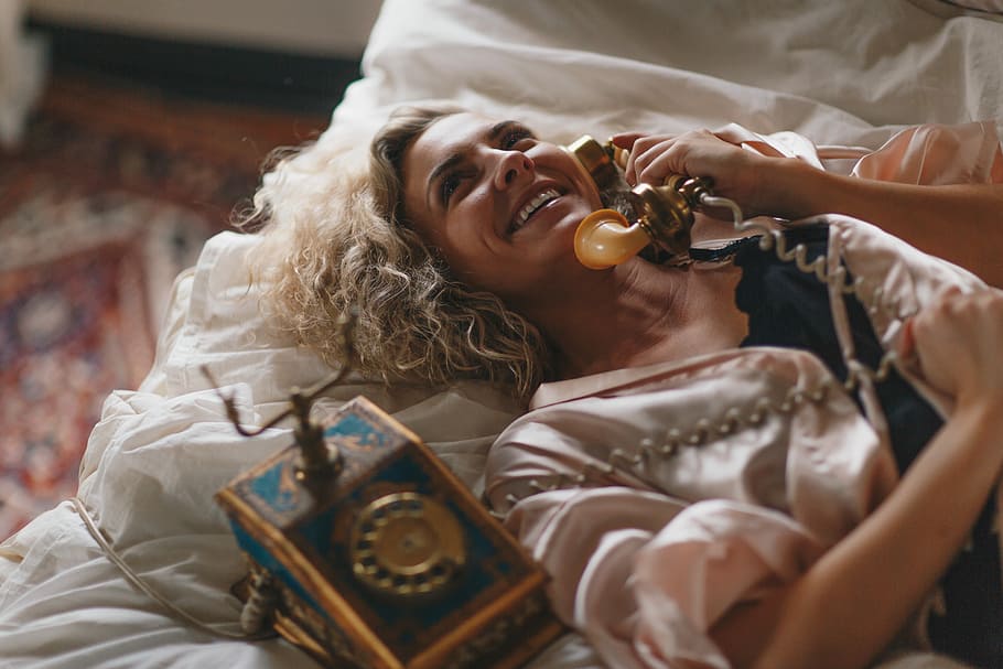 woman laying on white bed using a brown telephone, woman lying on bed holding rotary phone