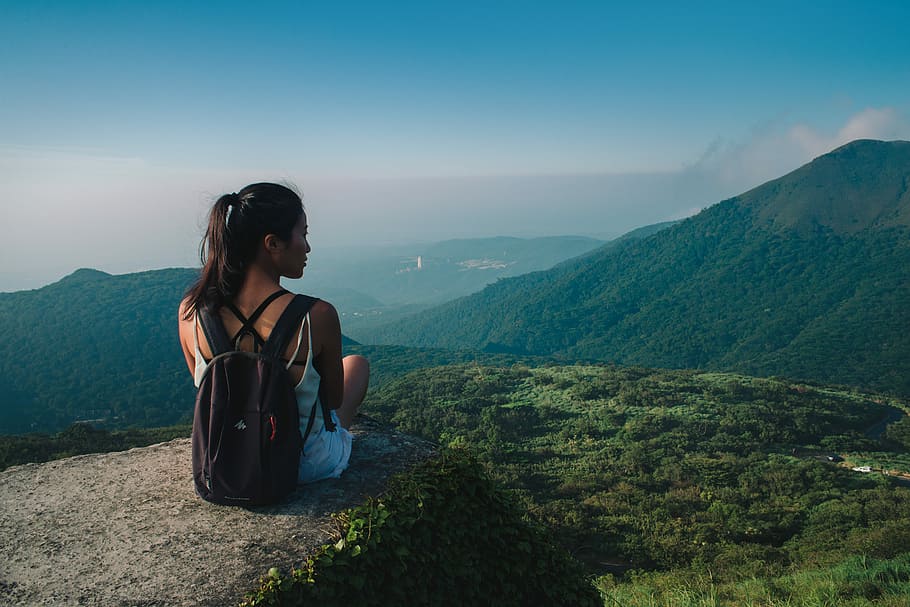 woman carrying black backpack sitting on top of hill, woman sitting on cliff with backpack facing mountain and trees
