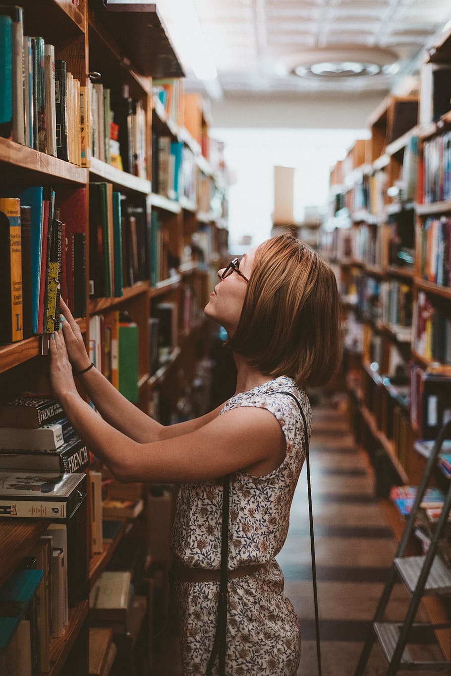woman standing between library book shelves, woman wearing white and brown floral dress standing near bookshelf looking up
