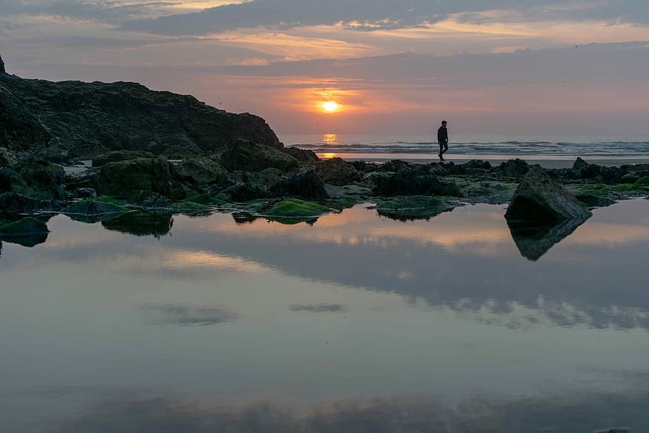 person walking beside body of water during sunset, person standing on rock near body of water HD wallpaper