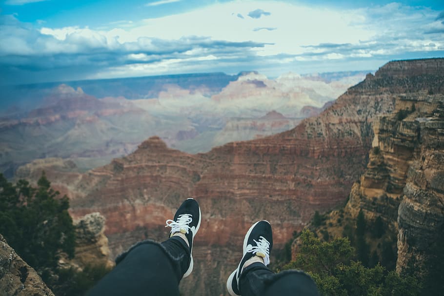 person sitting looking through mountain taken under white clouds during daytime, person wearing black shoes on hill over-viewing red and black mountains