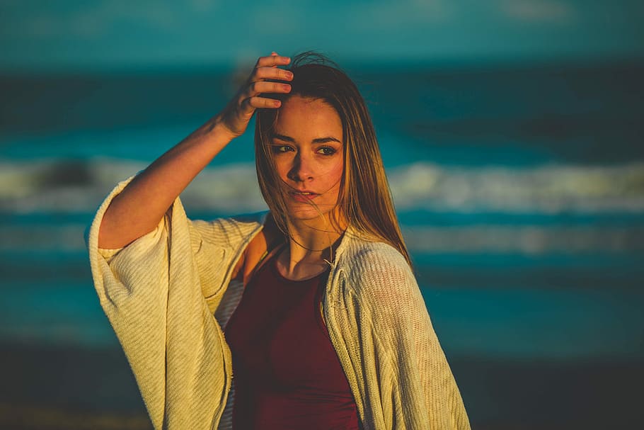 selective focus photography of woman in white cardigan standing near seashore holding her head, woman taking photo in front of seashore HD wallpaper
