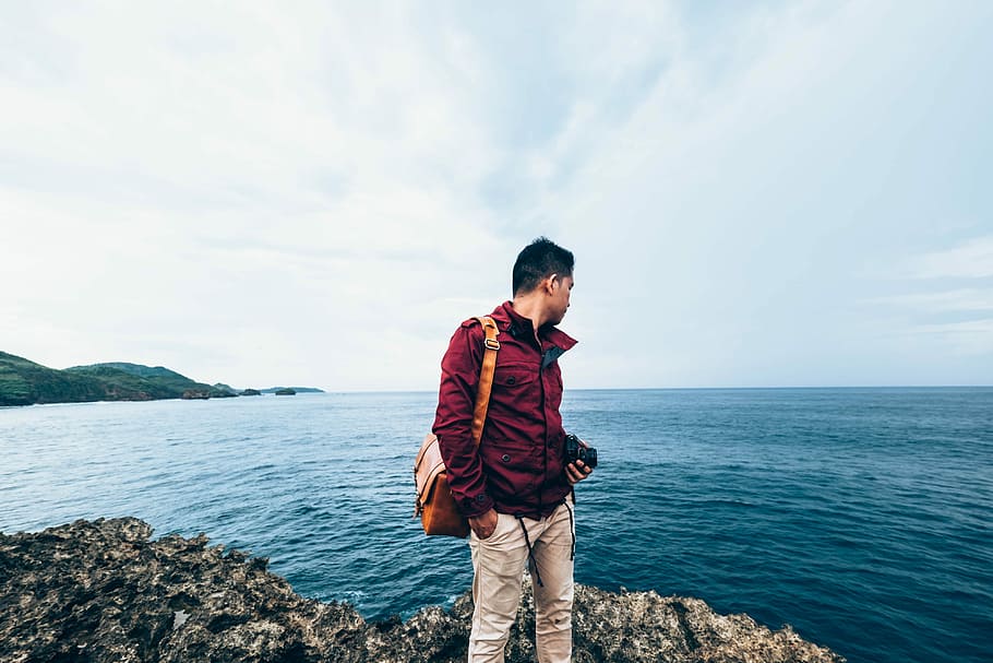 man wearing red zip-up jacket standing on top of rocky cliff beside body of water during daytime, man standing on a cliff near sea, HD wallpaper
