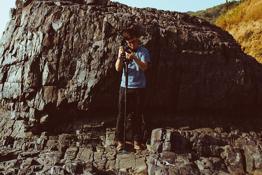 man holding camera while standing on cliff during daytime, man standing beside rock formation holding DSLR camera during daytime HD wallpaper