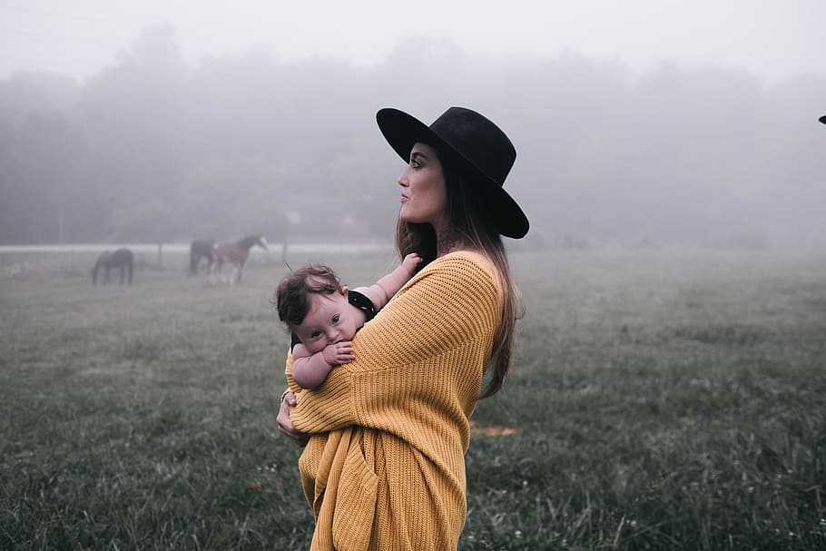 woman in yellow coat wearing black sun hat standing at green grass field while carrying baby, man carrying baby while fogging in fields