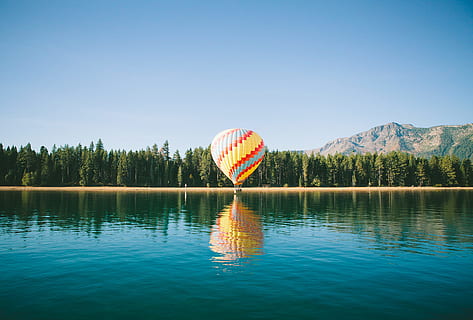 yellow, red, and blue hot air balloon near trees and body of water under blue sky during daytime, multicolored hot air balloon HD wallpaper