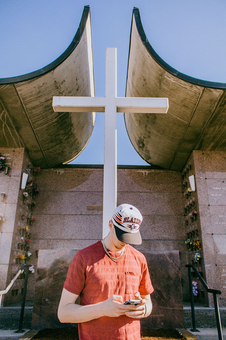man holding smartphone and standing near cross statue, man in orange crew-neck T-shirt using smartphone standing near wooden cross HD wallpaper