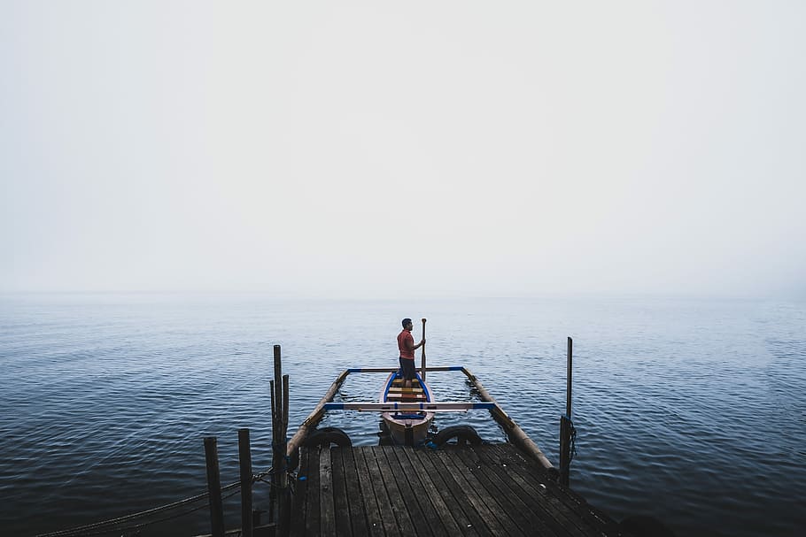 man standing on canoe near dock during daytime, man standing on beige row boat on body of water during daytime HD wallpaper
