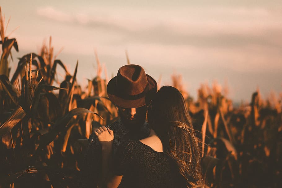 man and woman hugging each other in the field, man in brown fedora hat hugging woman in black top beside plants HD wallpaper