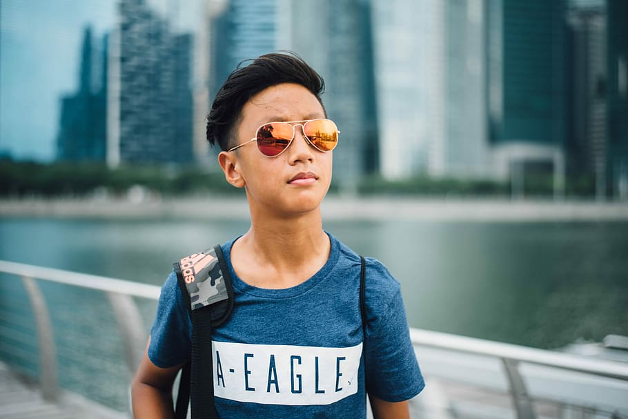 boy wearing yellow lens Aviator sunglasses near gray rail at daytime, boy wearing blue American Eagle crew-neck t-shirt and brown framed Aviator-style sunglasses carrying gray and black camouflage adidas bag