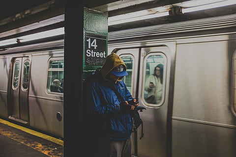 man in blue hoodie using smartphone in train station with train nearby, person wearing blue flat-brimmed cap and blue hoodie standing besides the pole HD wallpaper