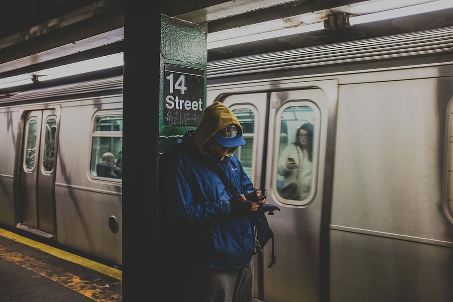 man in blue hoodie using smartphone in train station with train nearby, person wearing blue flat-brimmed cap and blue hoodie standing besides the pole HD wallpaper