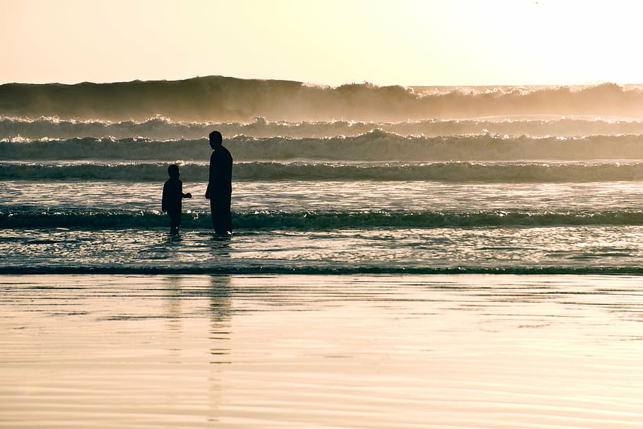 two person standing on seashore with sea waves during golden hour HD wallpaper