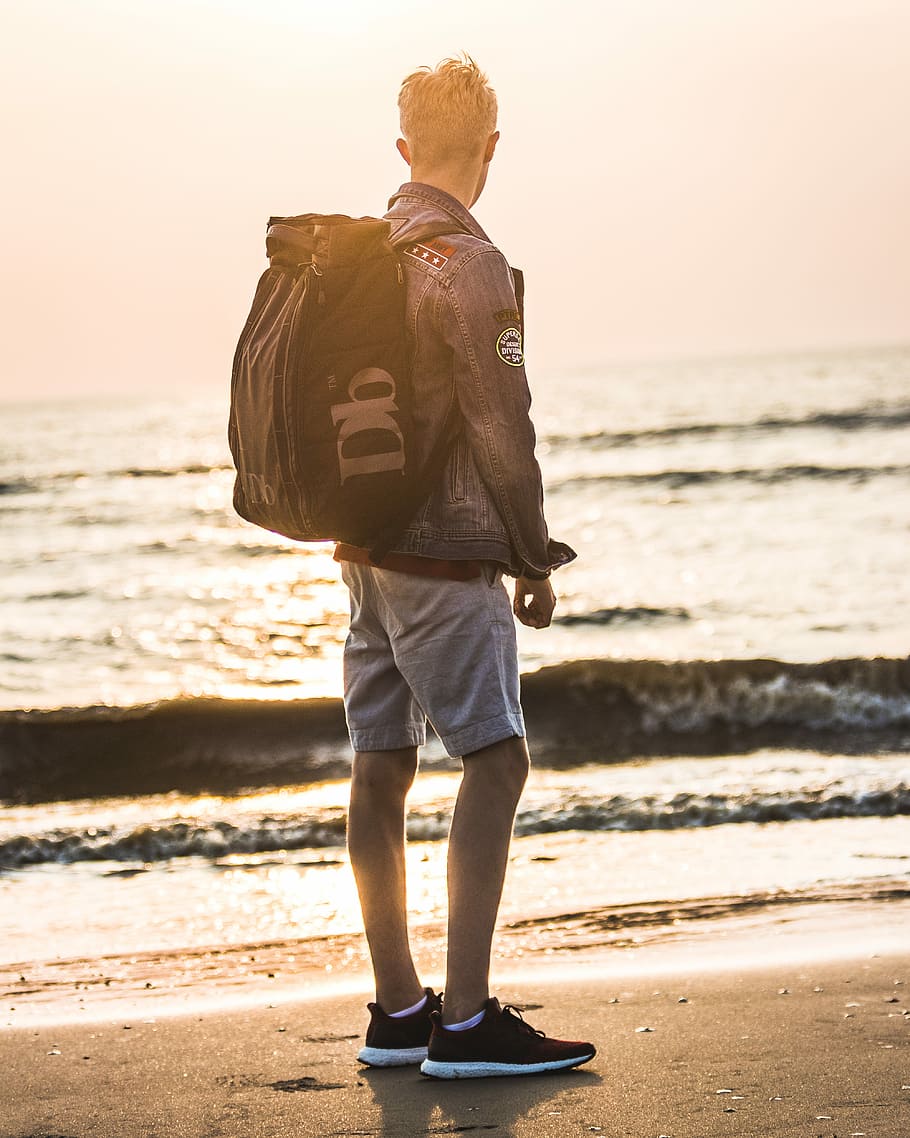 man carrying backpack standing near shoreline, man standing near shoreline
