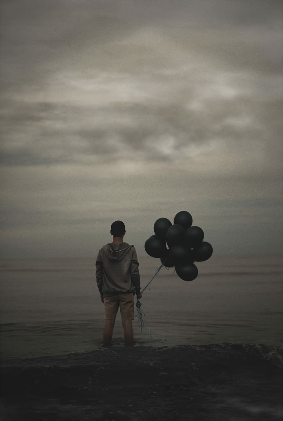 man holding balloons, man in gray hoodie standing on body of water while holding black balloons HD wallpaper