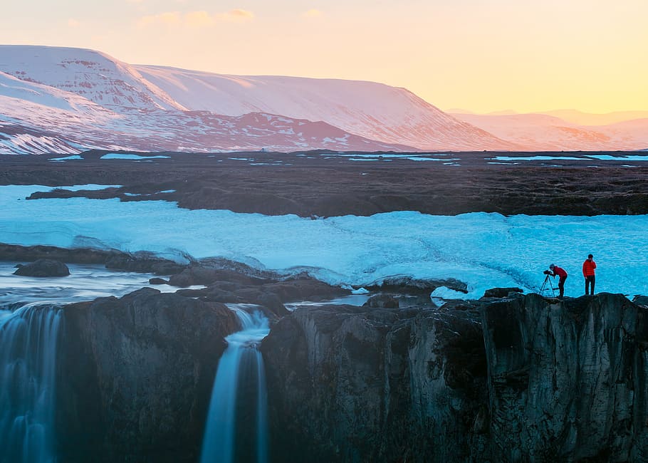 two persons standing on cliff with waterfall during daytime, photo of two person taking photo of snow covered mountain with waterfalls during daytime HD wallpaper