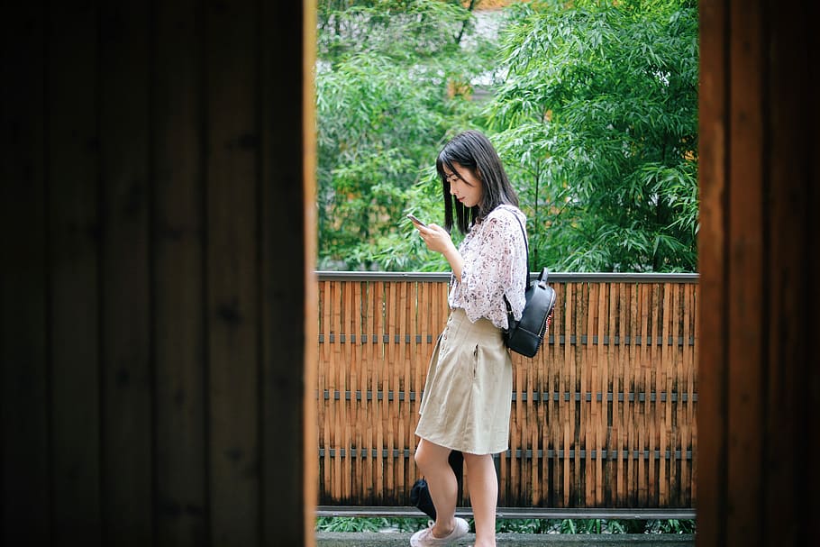 woman standing near brown and black wooden balcony over green leafed trees, woman using smartphone beside wall and trees during daytime HD wallpaper