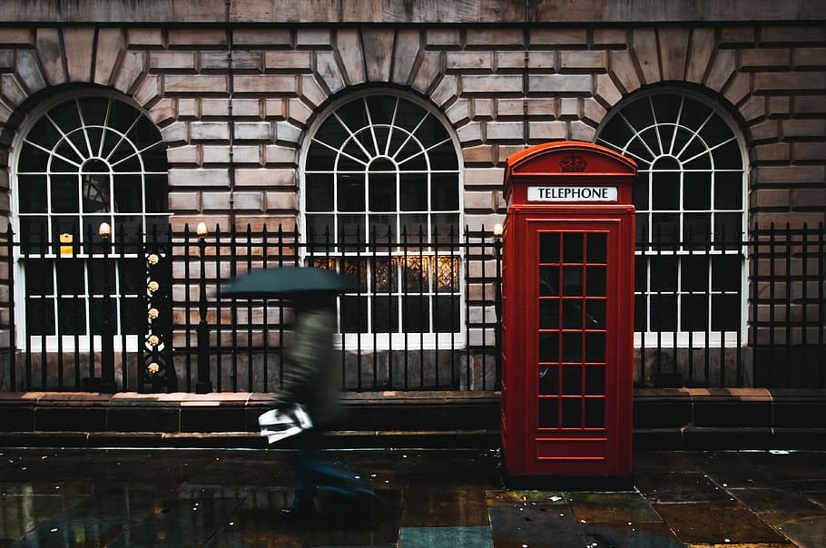 time lapse photography of woman walking on street while holding umbrella near London telephone booth beside wall, person walking beside red telephone book in front of black metal gate and brown brick building, HD wallpaper