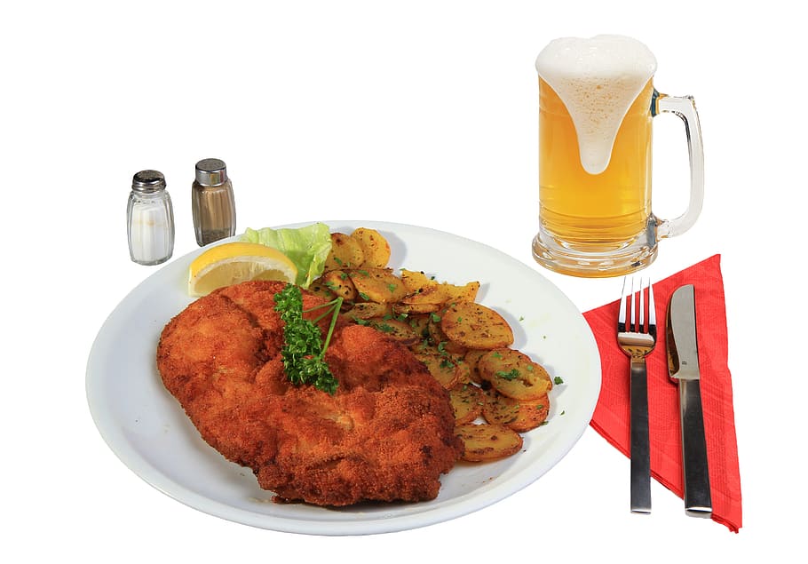 fried meat with sliced lemon on white plate beside condiment shakers, fork and knife, and beer mug filled with beer on white surface