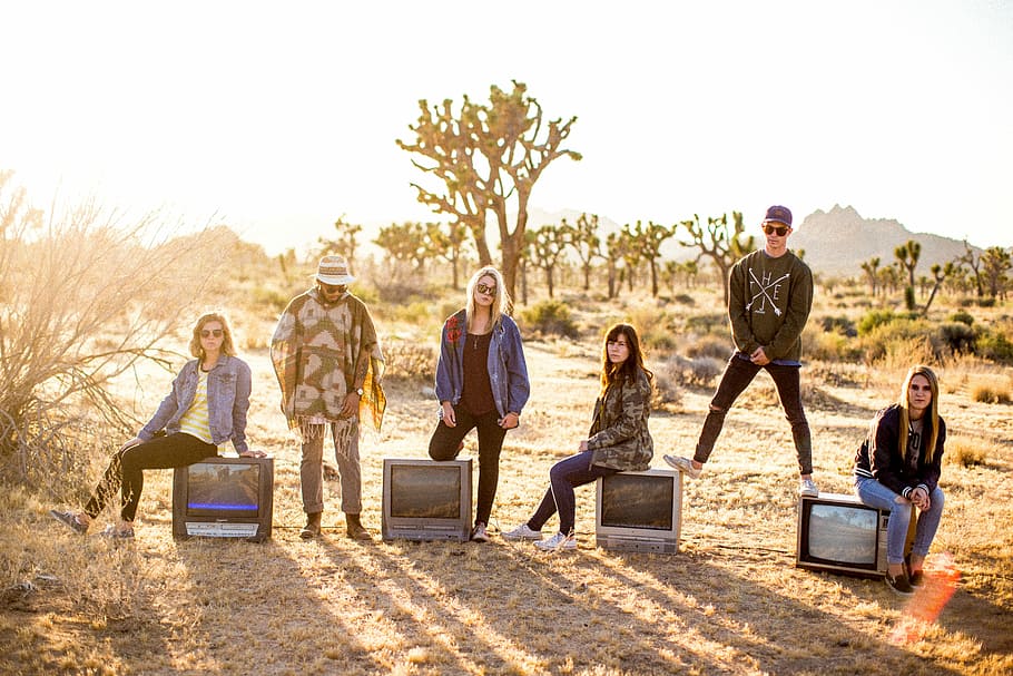 group of six people standing on desert, photograph of group of people seating on CRT televisions