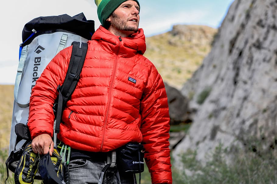 man wearing red bubble jacket and gray backpack standing beside rock HD wallpaper