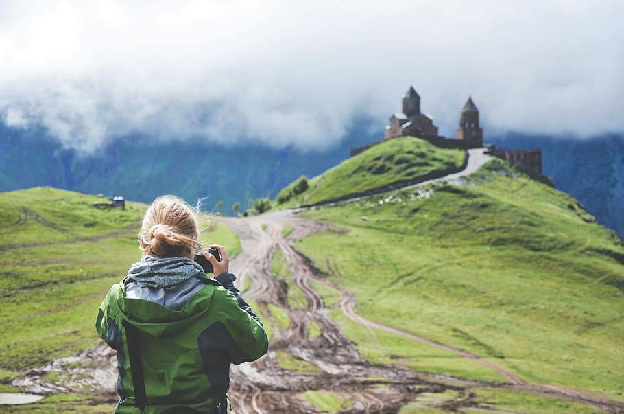 woman in green hooded windbreaker jacket holding dslr camera taking a photo of concrete building under white clouds during daytime HD wallpaper