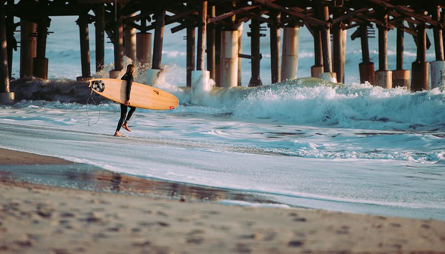 person holding surfboard standing on seashore in front of wave near wooden dock, walking man holding surfboard going to beach at daytime HD wallpaper