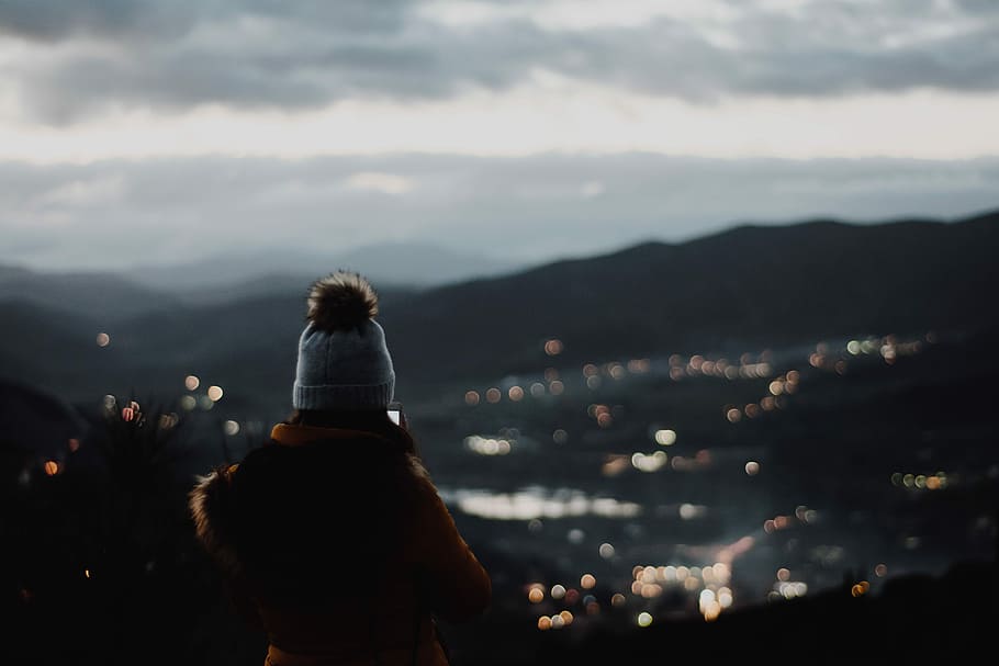 person standing on cliff with city lights as view, woman wearing white and brown bobble beanie looking at city