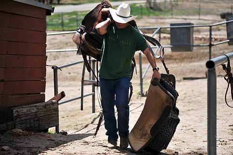 man in green crew-neck shirt holding brown horse saddle, cowboy HD wallpaper