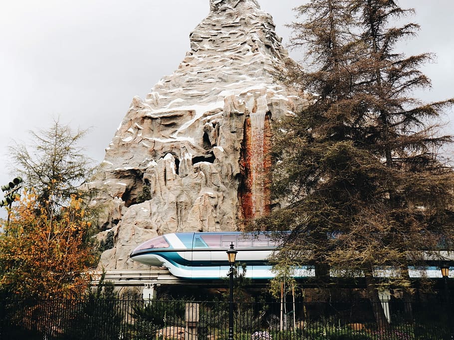 blue and white train beside brown rock mountain and pine trees, gray rock formation near green leaf tree HD wallpaper