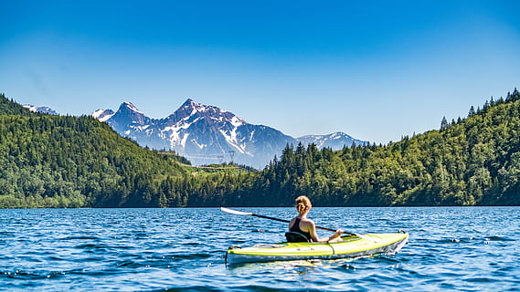 woman kayaking on lake during daytime, woman kayaking on body of water beside forest and mountains HD wallpaper