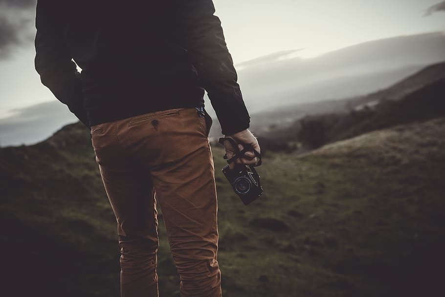 man standing uphill holding black digital camera, person standing on mountain slope holding camera HD wallpaper